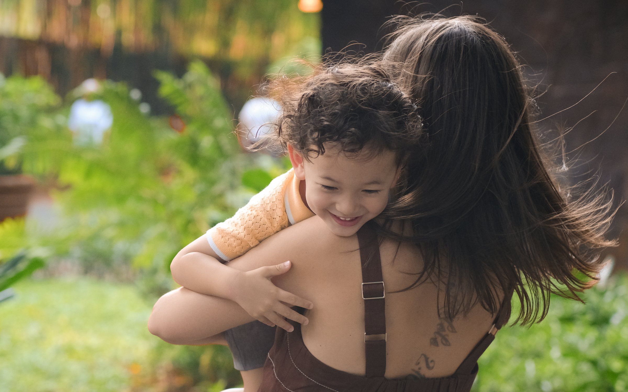 Curly-haired toddler hugging his mother from behind and grinning in a leafy garden in warm afternoon light