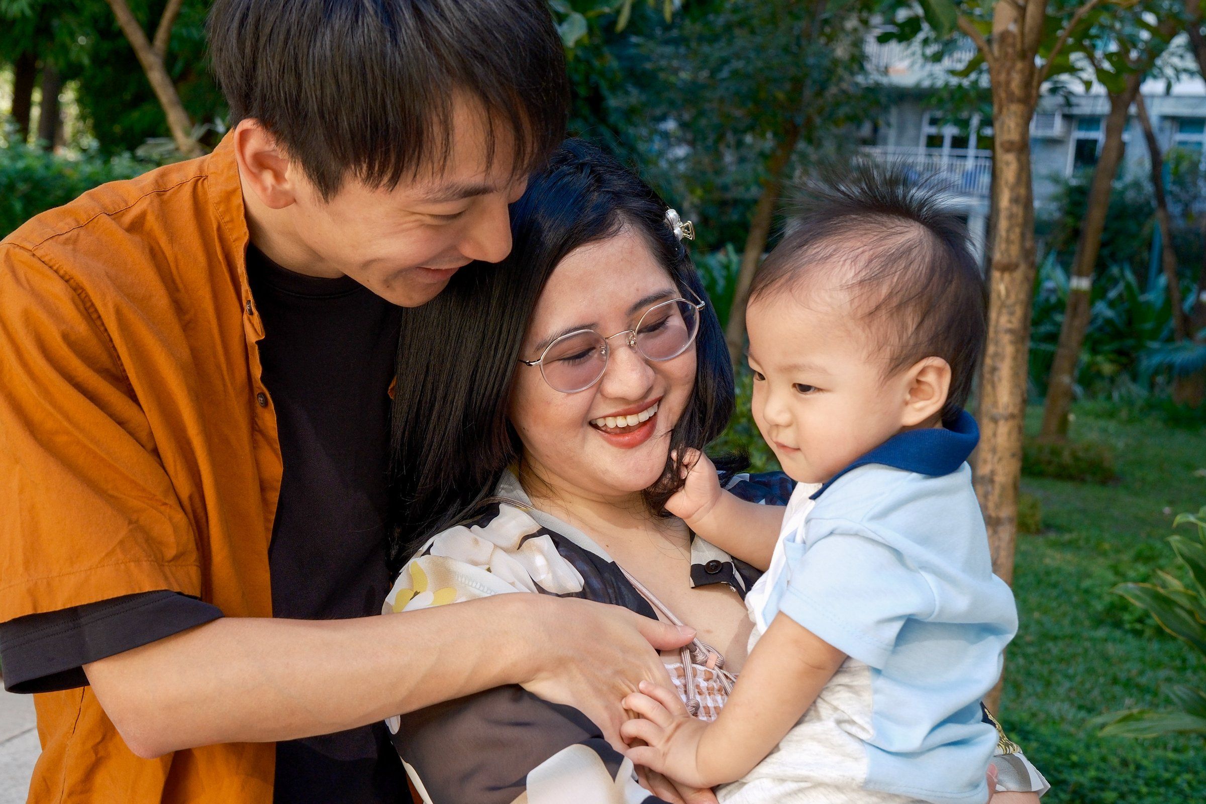 Parents laughing together while holding their baby son in a leafy park on a sunny afternoon