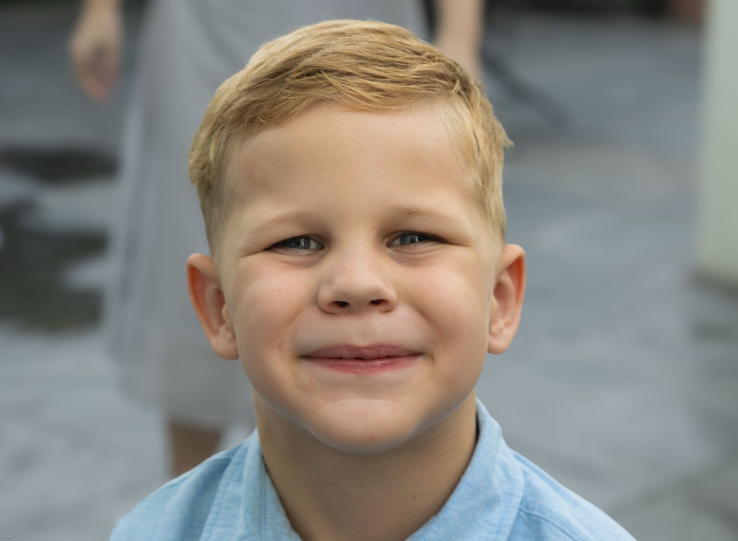 Close portrait of a smiling blond boy in a light blue shirt with a blurred courtyard behind